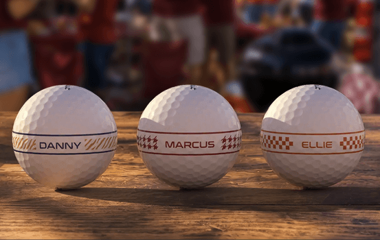 Three personalized golf balls with names and team-color AlignXL alignment patterns on a wooden tabletop.